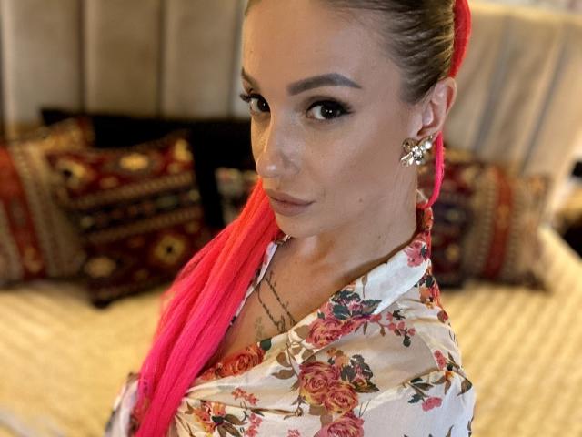 MelinFrosty with bright pink dreadlocks, floral shirt, and flower earrings, looking over her shoulder with a subtle smile, showing a small tattoo under her chin.