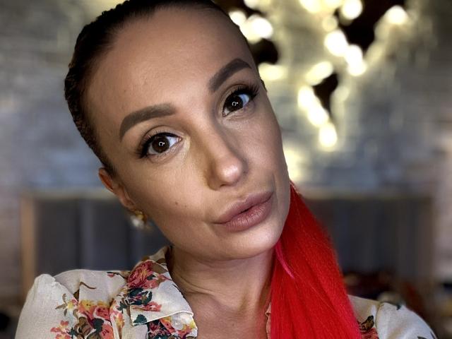 MelinFrosty with red ponytail, brown eyes, and floral shirt, looking at camera with slight smirk, showing off her smooth tits and cute face in selfie.