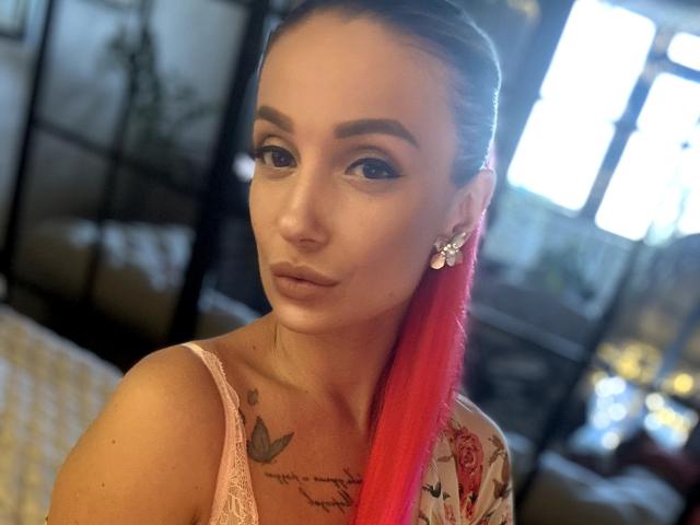 MelinFrosty with red ponytail, pink lace top, butterfly earrings, tattoos on chest, looking at camera, light brown skin, subtle makeup, pouty lips, medium-sized tits, soft light.