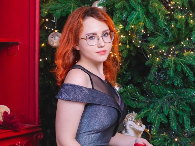 Teresa Birt mit roten Haaren, Brille und einem grauen, schulterfreien Kleid, hält eine kleine Statue und steht neben einem geschmückten Weihnachtsbaum. Ihre Brüste sind kaum sichtbar, ihr Hintern ist nicht zu sehen..