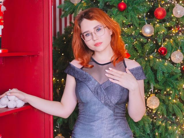 TeresaBirt with red hair, glasses, and gray dress, standing by a red shelf, touching her chest, holding a white ball, Christmas tree with ornaments in the background.