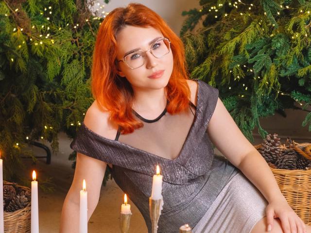 TeresaBirt with red hair, glasses, and grey off-shoulder dress, sitting among candles and greenery, showing her small tits and cute ass. Candle flames light her up.