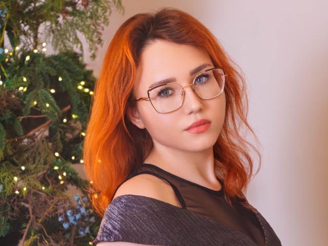 Teresa Birt, con cabello rojo, gafas y una expresión seria, vestida con una blusa negra transparente que deja ver sus hombros, frente a un árbol de Navidad con pequeñas luces amarillas..