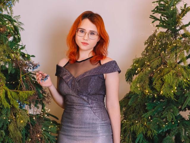 Teresa Birt mit roten Haaren, Brille und einem grauen, schulterfreien Kleid, steht zwischen zwei geschmückten Weihnachtsbäumen und hält einen Zweig in ihrer rechten Hand. Ihre Brüste sind klein..
