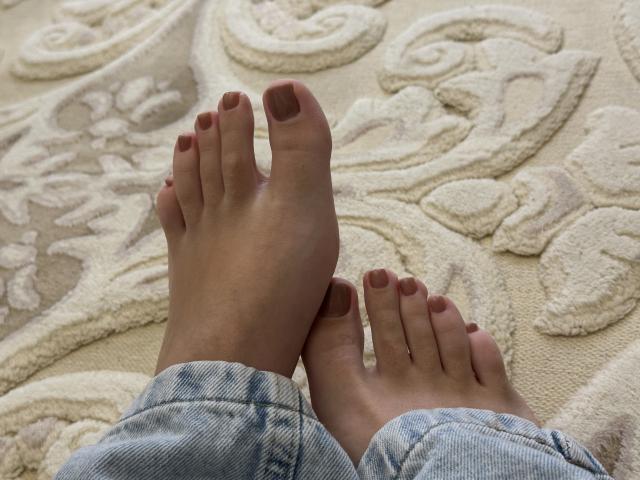 MargoFun's bare feet with brown-painted toenails, showing soft skin, and blue denim jeans, resting on a textured, cream-colored rug.