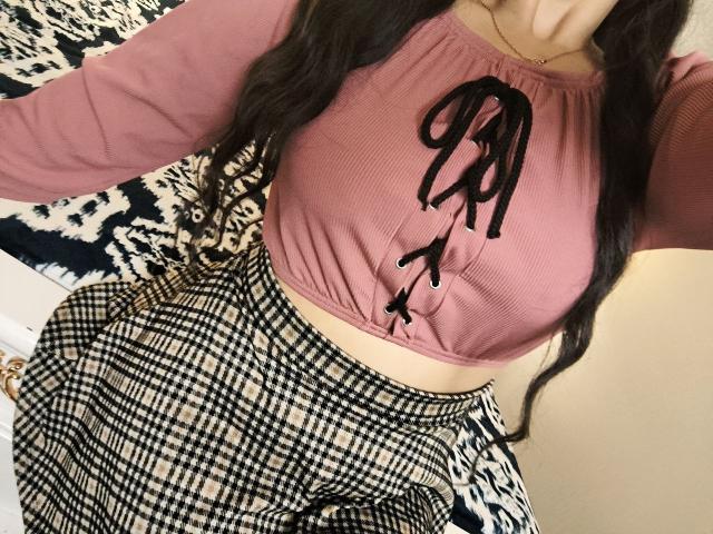 Niferttiti with large boobs, wearing a pink ribbed crop top with black lace-up, and a high-waisted black-and-white checkered skirt, long wavy brown hair.