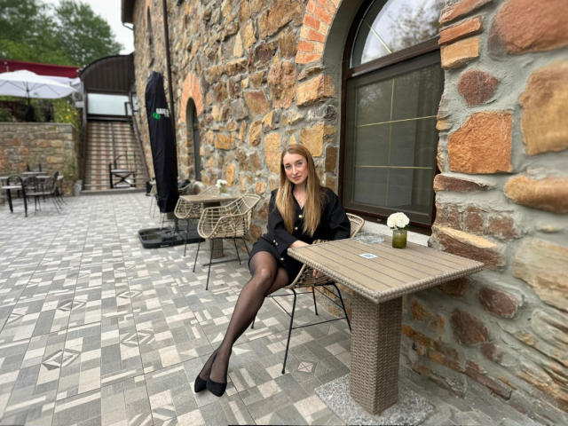 JamieSweetX, a blonde woman with long hair, sits at a wicker table against a stone wall with an arched window. She wears a black blazer, black shorts, and black high heels. Her legs are crossed, and she looks slightly to the right. The table has a small white flower in a glass vase and a card. The stone wall and floor pattern add texture. The background includes a white umbrella and more wicker furniture. The image is sharply focused.