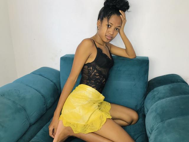 SaynahSisi with dark skin, small tits, in a black lace top and yellow lace skirt, sitting on a teal couch, hand in hair, looking at camera, smiling.