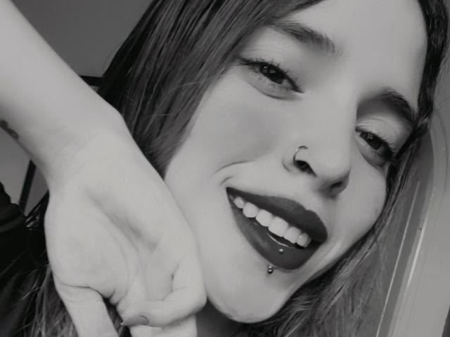 Black-and-white selfie of LovelyApril with dark lipstick, nose ring, and lip piercing, smiling with her hand near her chin, showing a septum piercing and a lower lip piercing.