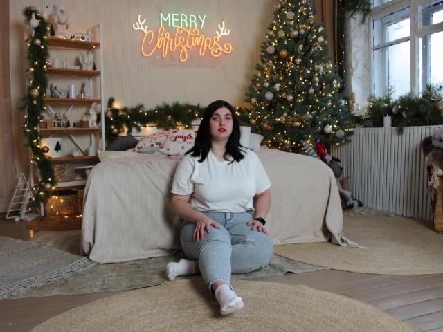 ScarletCargo, with black hair and red lipstick, sits on a rug, wearing a white shirt, grey pants, and white socks, in front of a Christmas tree with lights.