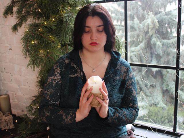 ScarletCargo with red lipstick, dark hair, lace green top, holding white candle in small basket, looking down, standing by window with green tree, soft yellow lights.