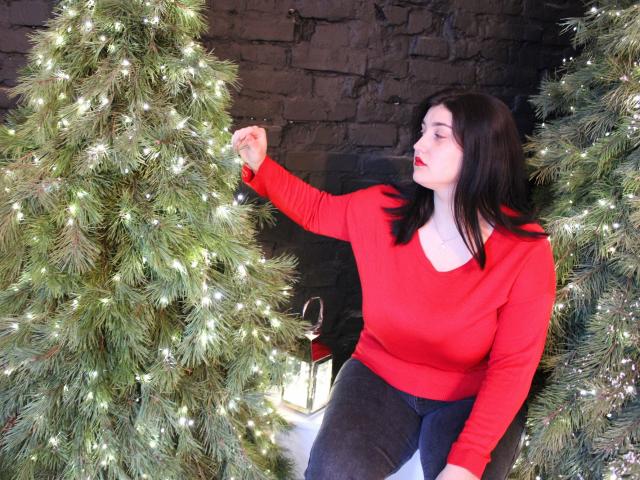 ScarletCargo, with black hair and red sweater, touches a Christmas tree's branch, standing between two lit trees, wearing black pants, looking to the side with red lipstick.
