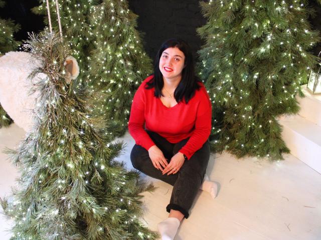 ScarletCargo with black hair, red sweater, black pants, and white socks, sitting between two green Christmas trees with white lights, smiling at camera. Her tits are small.