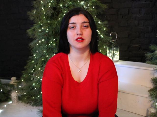 ScarletCargo with long black hair, red lipstick, and red long-sleeve top, wearing a necklace, sitting in front of a decorated Christmas tree with glowing lights.