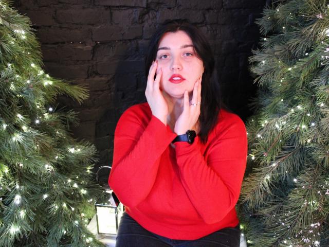 ScarletCargo with dark hair, red sweater, hands on cheeks, sitting between two Christmas trees with lights, looking at camera, black wristwatch, brick wall background, cute and attractive.