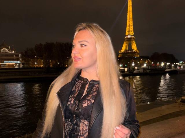 AmeliHot69 with long blonde hair, black lace top, black jacket, and cute smile, standing by a river with the Eiffel Tower glowing in the night background.