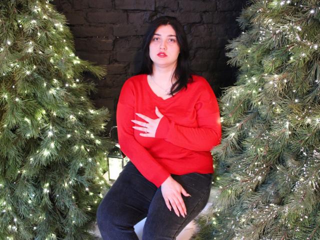 ScarletCargo with black hair, red sweater, and black pants, sitting between two lit-up Christmas trees, hand on chest, looking at camera, slightly parted red lips, medium tits.