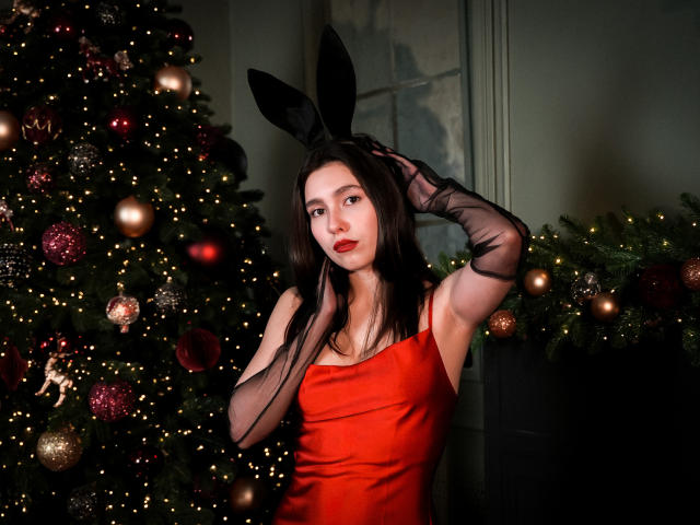 LounaLove with black bunny ears, red satin dress, and black sheer gloves, hands in her dark hair, standing next to a Christmas tree with red and gold ornaments.