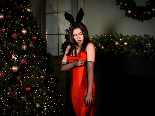 LounaLove with black bunny ears, red satin dress, and black gloves, standing by decorated Christmas trees, looking down with a slight frown, her tits partially visible.