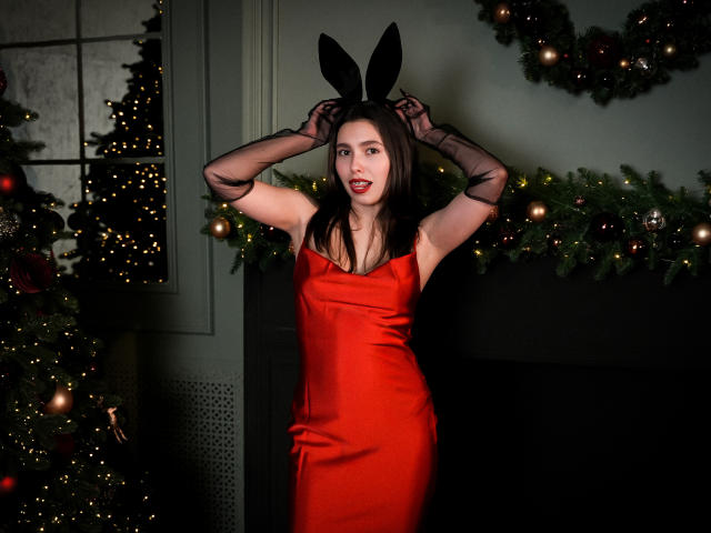 LounaLove in a red satin dress, black sheer gloves, and black bunny ears, with her hands up, showing off her tits and red lips, in front of Christmas trees.