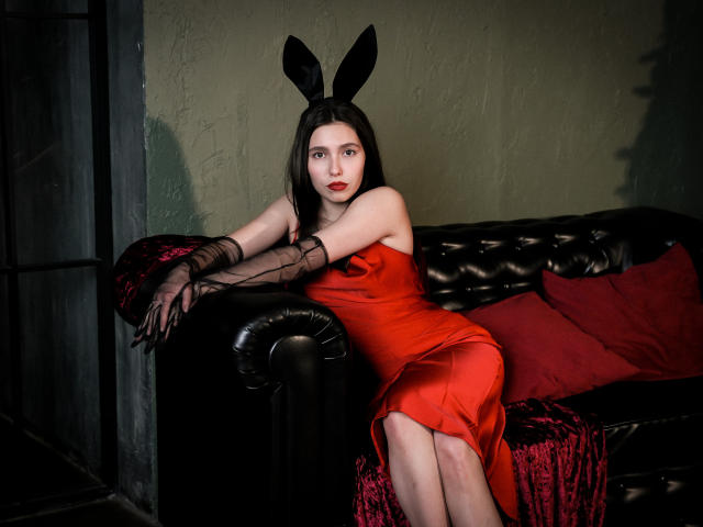 LounaLove with black bunny ears, red dress, and black mesh gloves, sitting on a black leather couch, red pillow, and red velvet blanket, showing her ass.