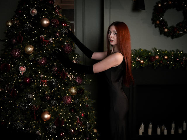 EllyCat with red hair, wearing a black dress and gloves, touching a Christmas tree adorned with red and gold ornaments, standing beside a dark fireplace with candles.