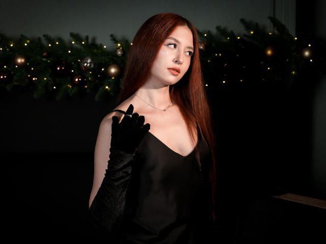 EllyCat with long red hair, wearing black satin dress and black velvet gloves, touching her chin, looking to the side, pale skin, subtle makeup, dark background with Christmas lights.