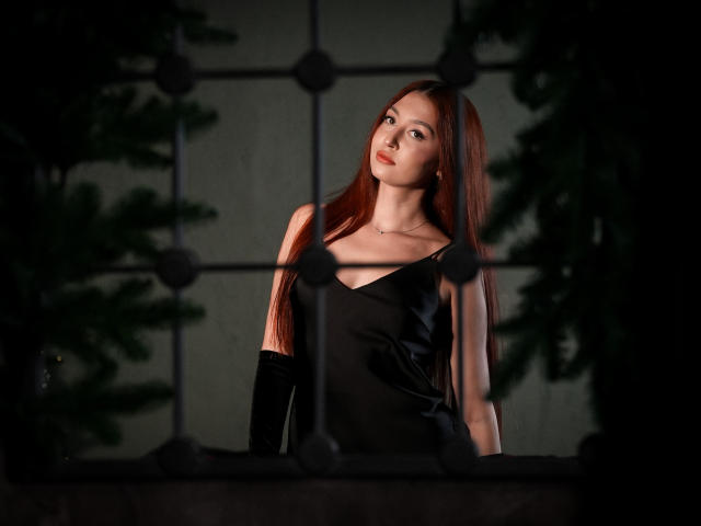 EllyCat with long red hair, black dress, and black gloves, standing in front of a dark, metal grid, looking slightly up with a calm expression. Boobs visible.
