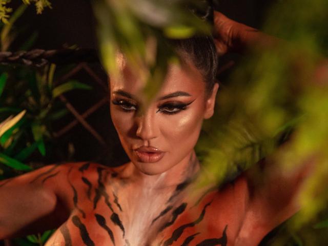 MellAnyass with tiger-striped tattoos, full lips, dark makeup, and raised arm, partially hidden by green foliage. Her tits are visible, and she has a sexy, intense expression.