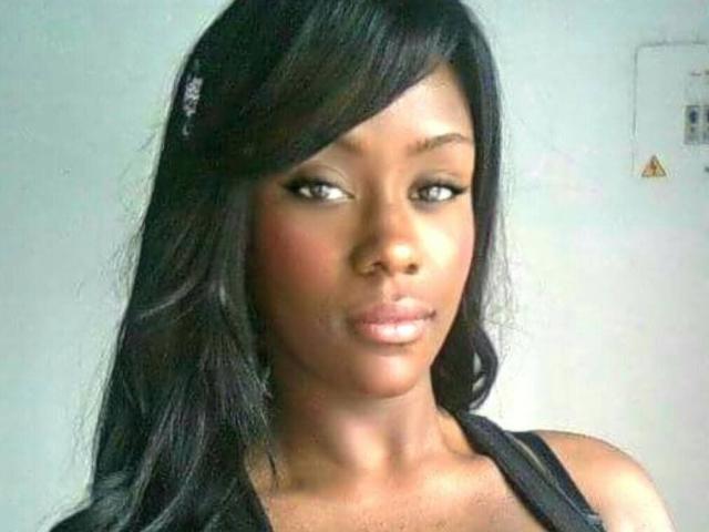 JhennyRousse has dark skin, long black hair, and is wearing a black top. She has full lips, arched eyebrows, and is looking to the side with a serious expression.