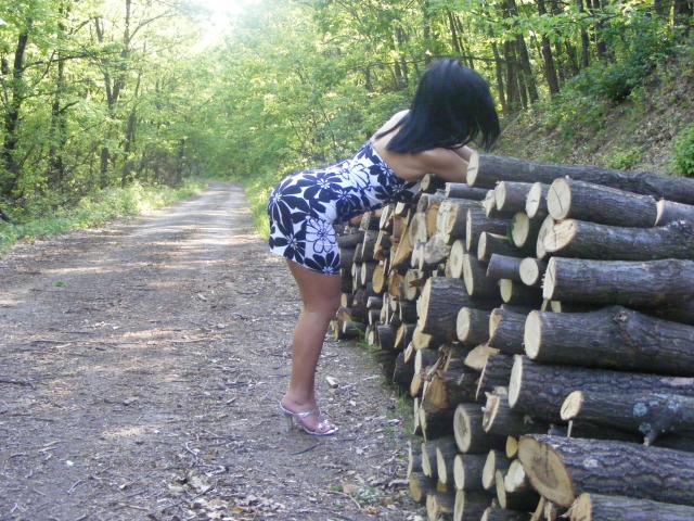 Gesucht: Eine Frau namens "Wantedcandy", in einem schwarz-weißen Blumenkleid, beugt sich vor und zeigt ihr Gesäß, während sie neben einem Stapel von Holzscheiten auf einem Waldweg steht..
