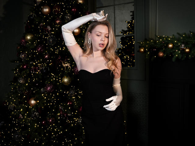 DellaLovely with long blonde hair, wearing a black strapless dress and white gloves, posing with her arm raised, showing off her tits and elegant earrings in front of a Christmas tree.