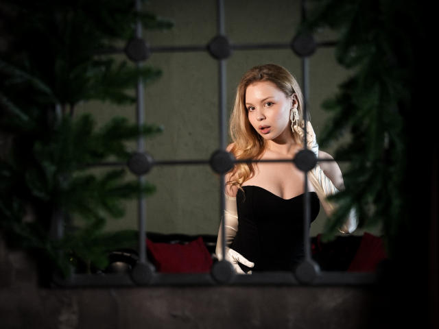 DellaLovely with blonde hair, wearing a black strapless dress, standing behind a metal fence, looking through it with her hand on her head, surrounded by dark greenery.