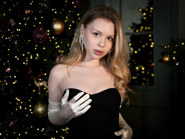 DellaLovely with blonde hair, wearing a black strapless dress, white gloves, and diamond earrings, covering her small tits with her hand, standing in front of a Christmas tree.