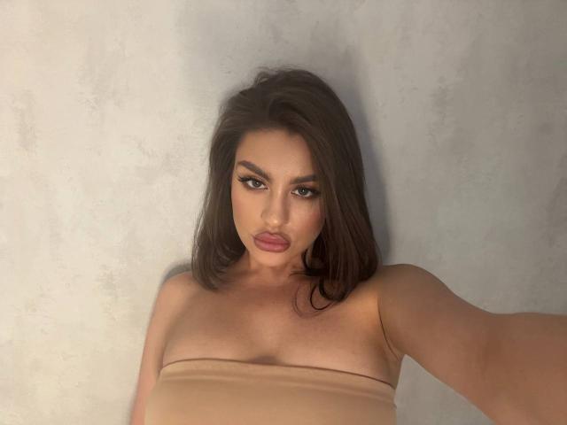 Close-up selfie of KristyDolly with medium-length, dark brown hair. She has thick, dark eyebrows, long, curled eyelashes, and full, pink lips. KristyDolly's brown eyes are slightly narrowed. She is wearing a strapless, beige top that exposes her shoulders and part of her upper chest. The texture of the background is rough and light gray. Her right arm is extended to hold the camera. The lighting is soft, highlighting her facial features. The overall composition is centered and focuses on her face and upper torso.