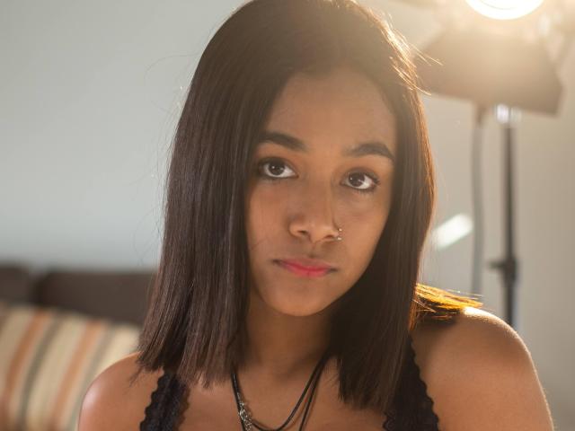 KamiBebe has medium brown skin, straight black shoulder-length hair, a nose ring, and wears a black lace top. Her brown eyes look slightly to the side. Soft lighting.