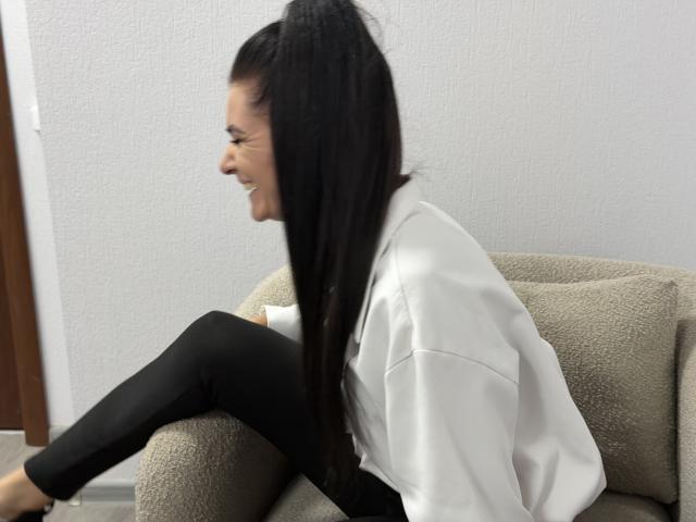 Viollia with long black hair in a ponytail, smiling, wearing a white shirt and black pants, sitting on a beige, textured armchair, showing her ass slightly.