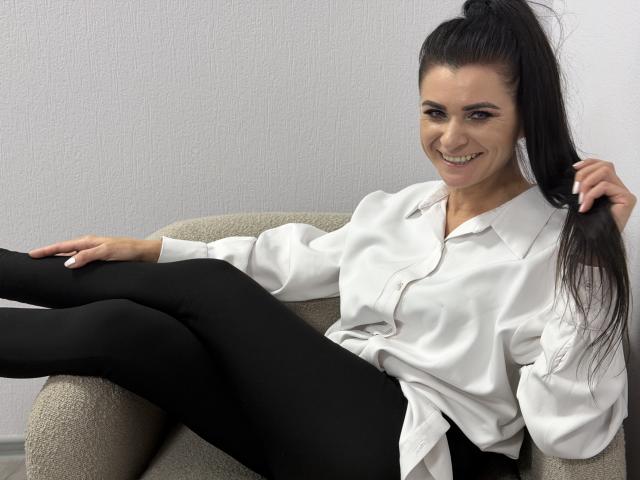 Viollia with long black hair in a ponytail, smiling, wearing a white shirt and black leggings, sitting on a beige chair, holding her hair with one hand.