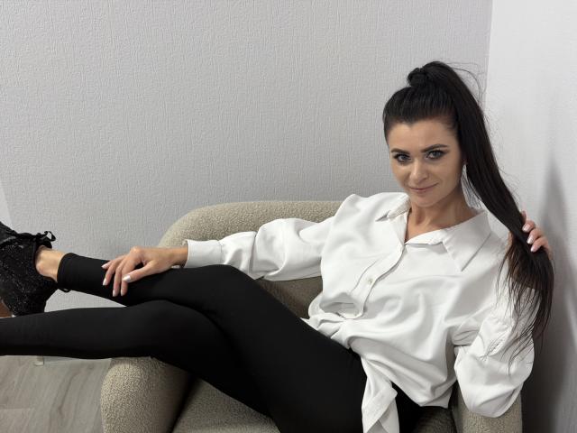 Viollia with long black ponytail, white button-up shirt, black pants, and black lace-up shoes, sitting in beige chair, smiling, legs crossed, right hand in hair, left on knee.
