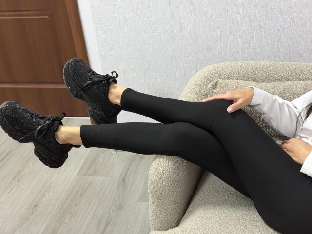 Viollia's legs are crossed and resting on a beige, textured armchair. She's wearing black, form-fitting leggings and black, glittery sneakers with laces. Her right hand is on the armchair, and her left arm is partially visible. The floor is light wood, and a brown door is in the background. The image is focused on her legs and shoes, with a slight upward angle. The texture of the armchair and leggings is smooth and shiny.