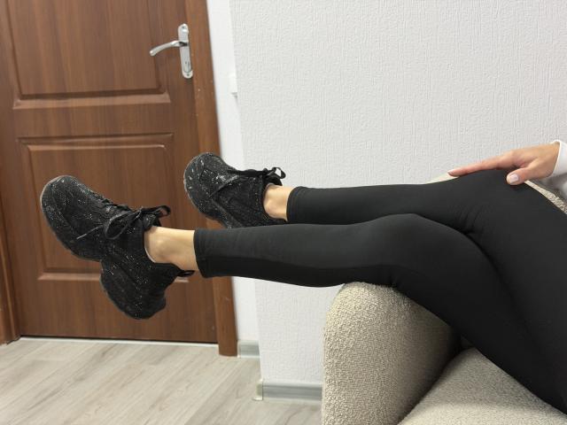 Viollia's black sparkly sneakers are on her black leggings-clad legs, resting on a beige chair. Her hand is on her right thigh. Wooden door in background.