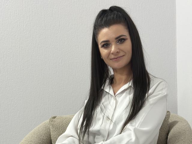 Viollia is a woman with long, straight black hair in a high ponytail. She's wearing a white button-up shirt and sitting on a beige, textured couch. Her light skin contrasts with her dark hair, and she has a subtle smile. Her eyebrows are thick and she has dark, defined eyes. The light in the image is even, highlighting her facial features and the texture of her shirt. The image is clear and focused on her upper body.