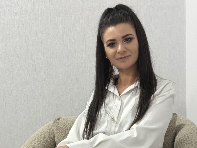 Viollia with long black hair in a ponytail, wearing a white button-up shirt, sitting on a beige couch, smiling slightly, arms crossed, brown eyes, light skin, subtle makeup.