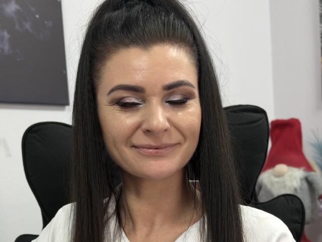 Viollia has long, straight black hair in a high ponytail. She has a light skin tone and a subtle smile. Her makeup includes shimmery, light brown eyeshadow and dark eyeliner. She's wearing a white shirt. The image is focused on her face, showing smooth, slightly shiny skin. The background has a black chair and a blurry red and white object. The overall composition is centered and close-up.