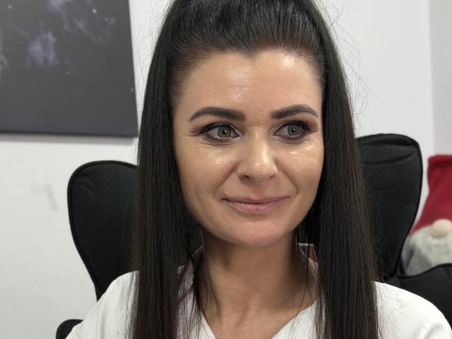 Viollia has shiny skin, green eyes, long dark hair in a high ponytail, and a subtle smile. She's wearing a white shirt, sitting in a black chair.