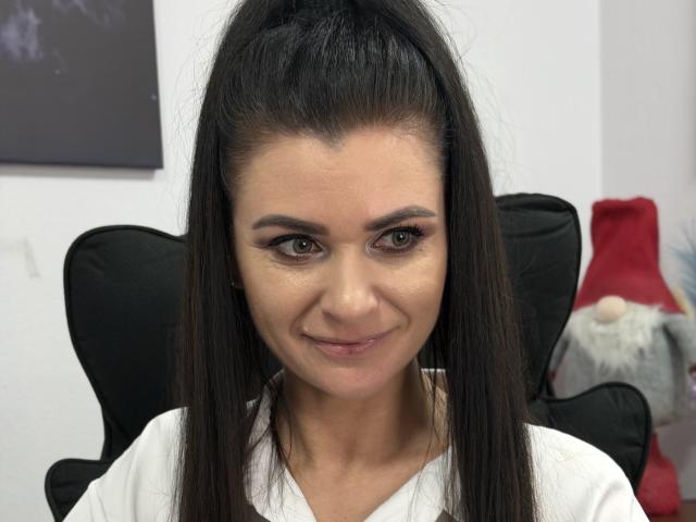 Viollia with straight black hair in a high ponytail, brown eyes, subtle makeup, and a slight smile, wearing a white shirt, sitting in a black chair.