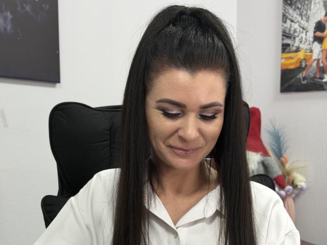 Viollia with long black hair in a high ponytail, wearing a white shirt, has subtle makeup, looking down with a slight smile, sitting in a black office chair.