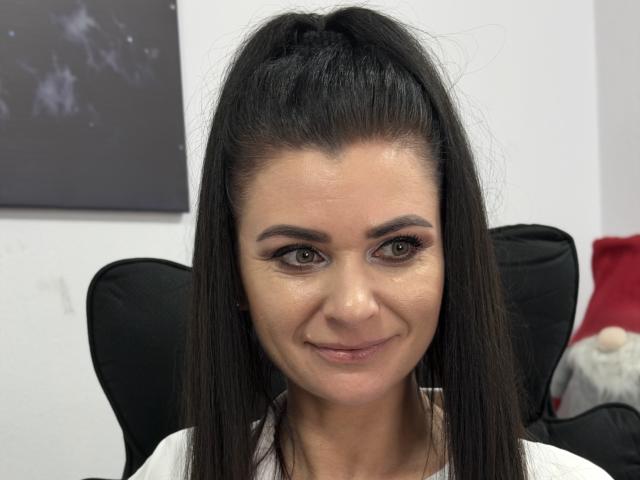 Viollia with long black ponytail, brown eyes, subtle smile, wearing white top, sitting in black chair, slightly glossy skin, minimal makeup, looking directly at camera.