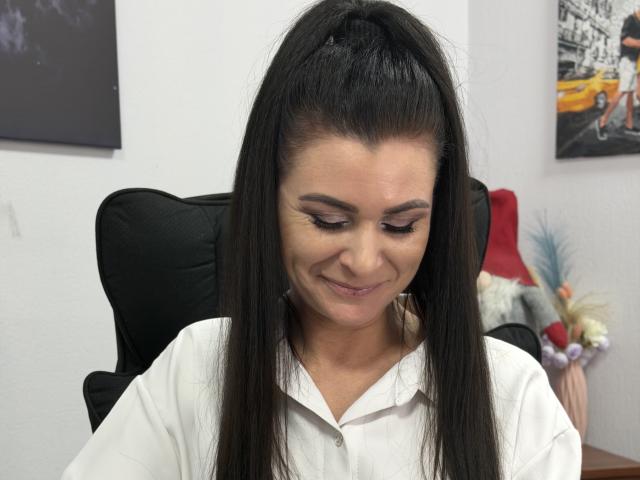 Viollia has long, straight black hair in a high ponytail. She's wearing a white polo shirt and has subtle, natural makeup with dark eyeliner. She's looking down with a slight smile. The background includes a black chair, a red lamp, and a blurred cityscape photo. The image has a watermark in the bottom right corner. The focus is on Viollia's face and hair. The colors are mostly neutral with a few bright accents.