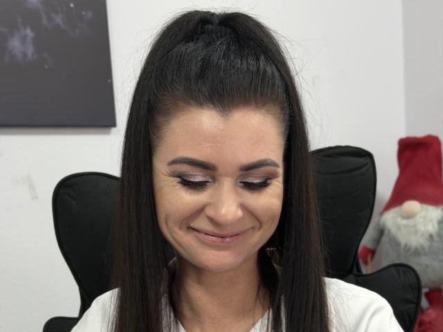 Viollia has straight, dark brown hair in a high ponytail. She's smiling slightly with closed eyes, showing subtle makeup with pink eyeshadow and black eyeliner. Her skin is light olive. She's wearing a white top. The background has a black chair and a red stuffed animal. The image is sharp, with clear details in her hair and face. The overall style is realistic.